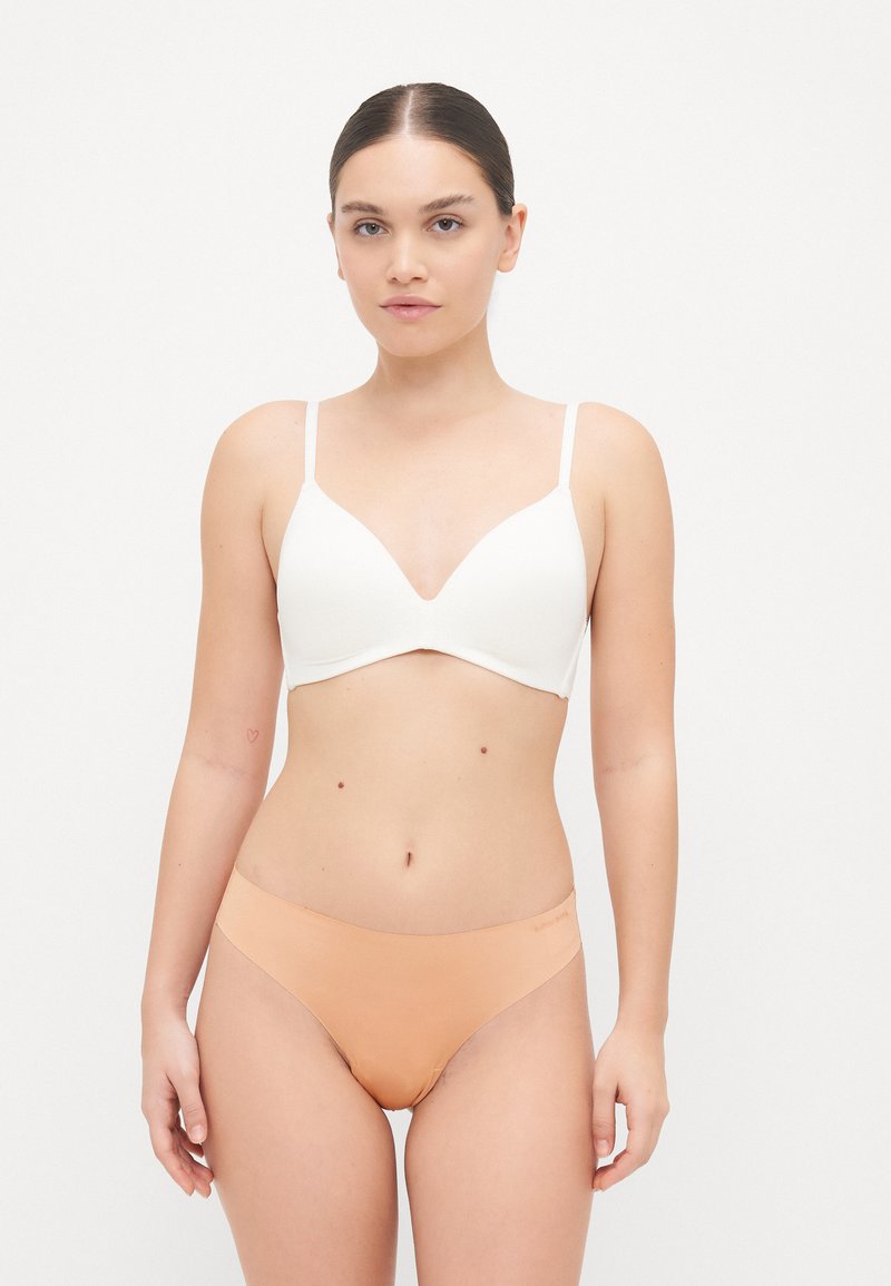 Weißer nahtloser BH mit verstellbaren Trägern und V-förmigem Ausschnitt. Hellpfirsichfarbene nahtlose Panties mit glatter Oberfläche und minimaler Detailgestaltung.