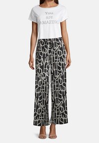 Žena v bílém zkráceném tričku s nápisem "Jsi ÚŽASNÁ" a černých širokých kalhotách s velkými abstraktními bílými písmeny.