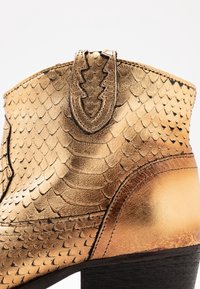 Golden textured ankle boot with a scaly pattern, smooth leather accents, a decorative stitched pull tab, and a black wooden heel.