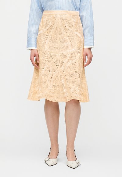 Femme portant une jupe crochetée beige mi-longue, un chemisier à manches longues rayé bleu et des chaussures blanches pointues avec des nœuds.