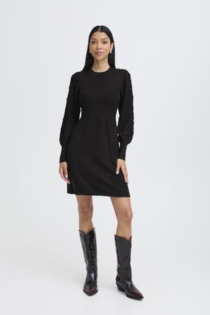 Robe noire tricotée avec des manches longues texturées, un corsage ajusté et une jupe évasée. Associée à des bottes de cowboy noires brillantes.