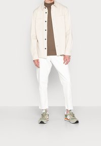 Light beige button-up shirt, brown sweater, white pants, and green sneakers with orange accents. Notable fabric textures and slim-fit design.