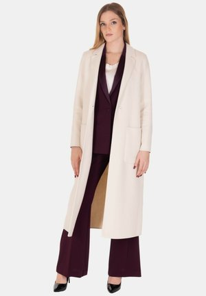 Lungo cappotto color crema realizzato in tessuto morbido, con taglio dritto, due tasche frontali e rever a notched, indossato sopra un abito bordeaux.