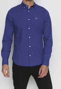 Camisa azul de botones con un pequeño patrón geométrico blanco. Botones blancos y un cuello con un logo de la marca en el pecho. Mangas largas.
