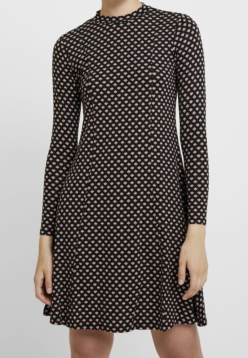 Långärmad åtsittande klänning med svart bakgrund och små beige geometriska diamantmönster, bärs av en stående person med avslappnade händer vid sidorna.