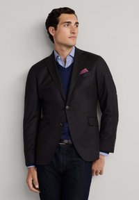 Blazer scuro con due tasche frontali, indossato sopra un maglione blu navy e una camicia azzurra chiara. Notevole fazzoletto da taschino con motivo rosa. Jeans scuri.