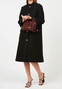 Femme portant un manteau midi noir avec des boutons dorés, tenant un sac à main en cuir marron et portant des sandales à talons hauts noires à lanières.