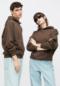 Braune Kapuzenpullover mit Kängurutaschen, kombiniert mit hellblauen, weiten Jeans. Eine Person trägt schwarze Sonnenbrillen. Einfacher, lockerer Schnitt.