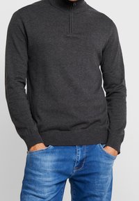 Dunkelgrauer Pullover mit hohem Kragen und halbem Reißverschluss, aus einem glatten Strickmaterial hergestellt. Ribbelbündchen und Saum. Über blauen Jeans getragen.