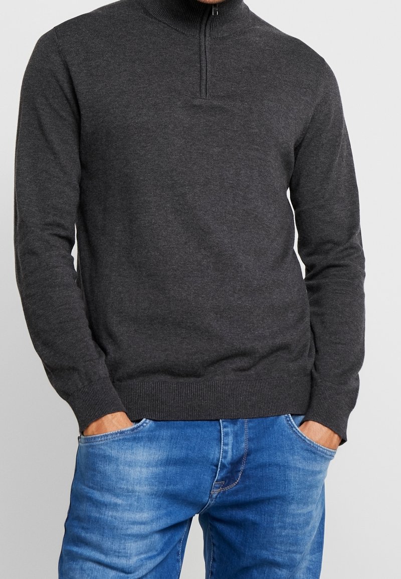 Dunkelgrauer Pullover mit hohem Kragen und halbem Reißverschluss, aus einem glatten Strickmaterial hergestellt. Ribbelbündchen und Saum. Über blauen Jeans getragen.