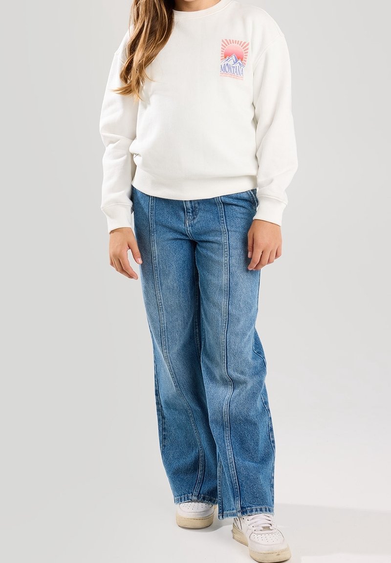 Enfant portant un sweat-shirt blanc avec un motif Montana, un jean bleu à jambes larges et des baskets blanches, debout devant un fond uni.