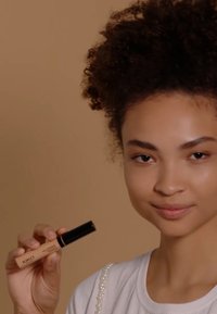 Correcteur KIKO avec un tube beige et un bouchon noir tenu par une personne, sur un fond neutre ; la personne porte une chemise blanche avec des accents de perles.