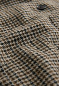 Vævet stof med et houndstooth-mønster i beige og brun, med sorte accenter. En sort knap er synlig på overfladen.