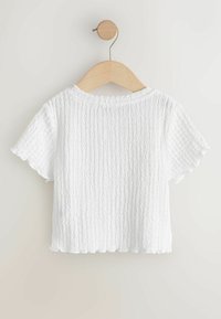 Witte korte mouwen top met een gestructureerde, gekreukte stof. Heeft een ronde hals en een gegolfde zoom, hangend aan een houten hanger tegen een lichte achtergrond.