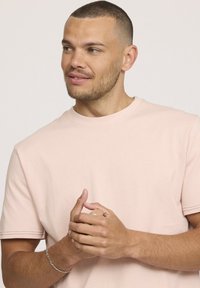 T-shirt rose pâle à manches courtes en tissu doux, avec un col rond et des détails de couture subtils. Les mains du mannequin sont jointes.