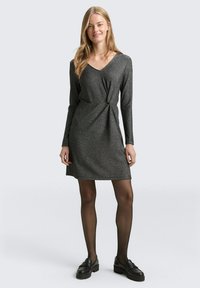 Schwarzes Langarmkleid mit leichtem Schimmer, V-Ausschnitt und einer Knoten-Detailung an der Taille, kombiniert mit schwarzen Loafers und Strumpfhosen.
