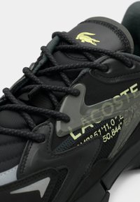 Black Lacoste sneaker with dark laces, yellow crocodile logo on tongue, and yellow coordinates printed near the brand name on the side.