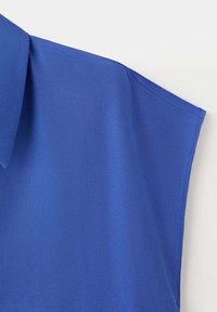 Blouse bleue à manches courtes avec un col plié, tissu lisse et coutures propres sur un fond clair.