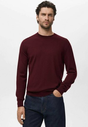 Burgundy crew neck sweater made of soft knit fabric, with long sleeves and ribbed cuffs. Paired with dark jeans, standing against a light background.