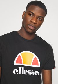 Camiseta negra de algodón, con un gran gráfico colorido en amarillo, rojo y blanco, con el nombre de la marca "ellesse" debajo del diseño.