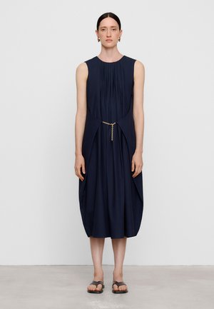 Vrouw met donker haar in een mouwloze donkerblauwe jurk met plooien en een gouden kettingriem, donkergekleurde sandalen dragend, staand tegen een effen witte achtergrond.