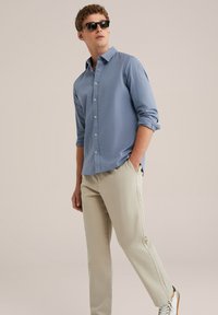 Cămașă albastră deschis cu mâneci lungi, asociată cu pantaloni chino bej și adidași albi, purtată de un bărbat care poartă ochelari de soare.