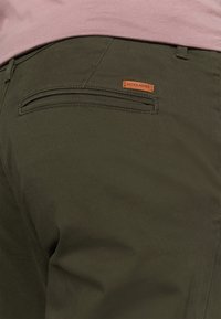 Tummanvihreät housut pehmeästä kankaasta, jossa on sileä pinta. Takana on tasku ja vyötäröllä ruskea logonauhainen etiketti.