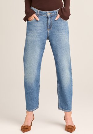 Frau trägt hellblaue High-Waist-Jeans mit geradem Bein, Hände in den Taschen und braune Absatzschuhe vor neutralem Hintergrund.