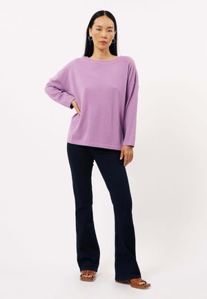 Haut à manches longues violet avec une coupe décontractée, associé à un jean évasé noir. Des sandales plates notables complètent le look. Fond blanc uni.