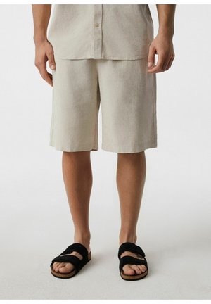 Mężczyzna w beżowych krótkich spodenkach do kolan, pasującej beżowej koszuli i czarnych otwartych sandałach stoi na białej podłodze.