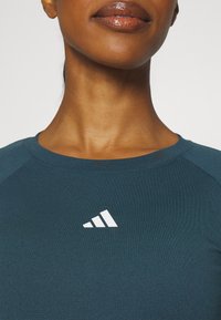 Grön atletisk topp med en slät textur, med en vit logotyp av tre diagonala ränder på bröstet och en rundad halsringning.