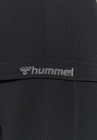 Svarta sportleggings med en slät textur och räfflade accenter. Har en grå "hummel"-logga på midjebandet.