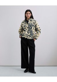 Chaqueta acolchada en crema y azul marino con un patrón de hojas. Presenta un cuello alzado, cierre de cremallera y dos bolsillos laterales. Combinada con pantalones de pierna ancha negros.