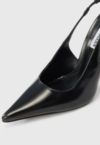 Μαύρο πέδιλο από λουστρίνι με ψηλό τακούνι, μυτερή μύτη και ανοιχτή πλευρική σχεδίαση, με γυαλιστερό φινίρισμα και μινιμαλιστικό στυλ.