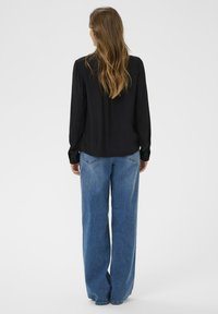 Kvinde med langt brunt hår iført en sort bluse med lange ærmer og løse blå jeans, stående vendt væk på en ensfarvet baggrund.