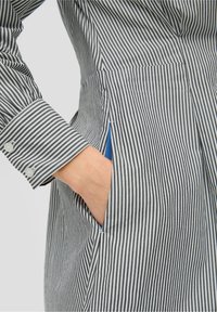 Chemise rayée avec des lignes verticales noires et blanches, en coton, col classique, poignets à boutons, poche latérale avec accent bleu.