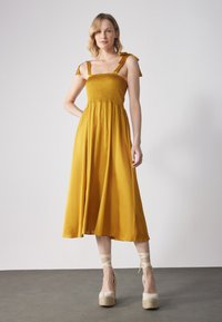 Sennepsgul kjole med smocket liv, justerbare knyteseler og en flytende A-linje skjørt, kombinert med kremfargede espadrille-wedges.