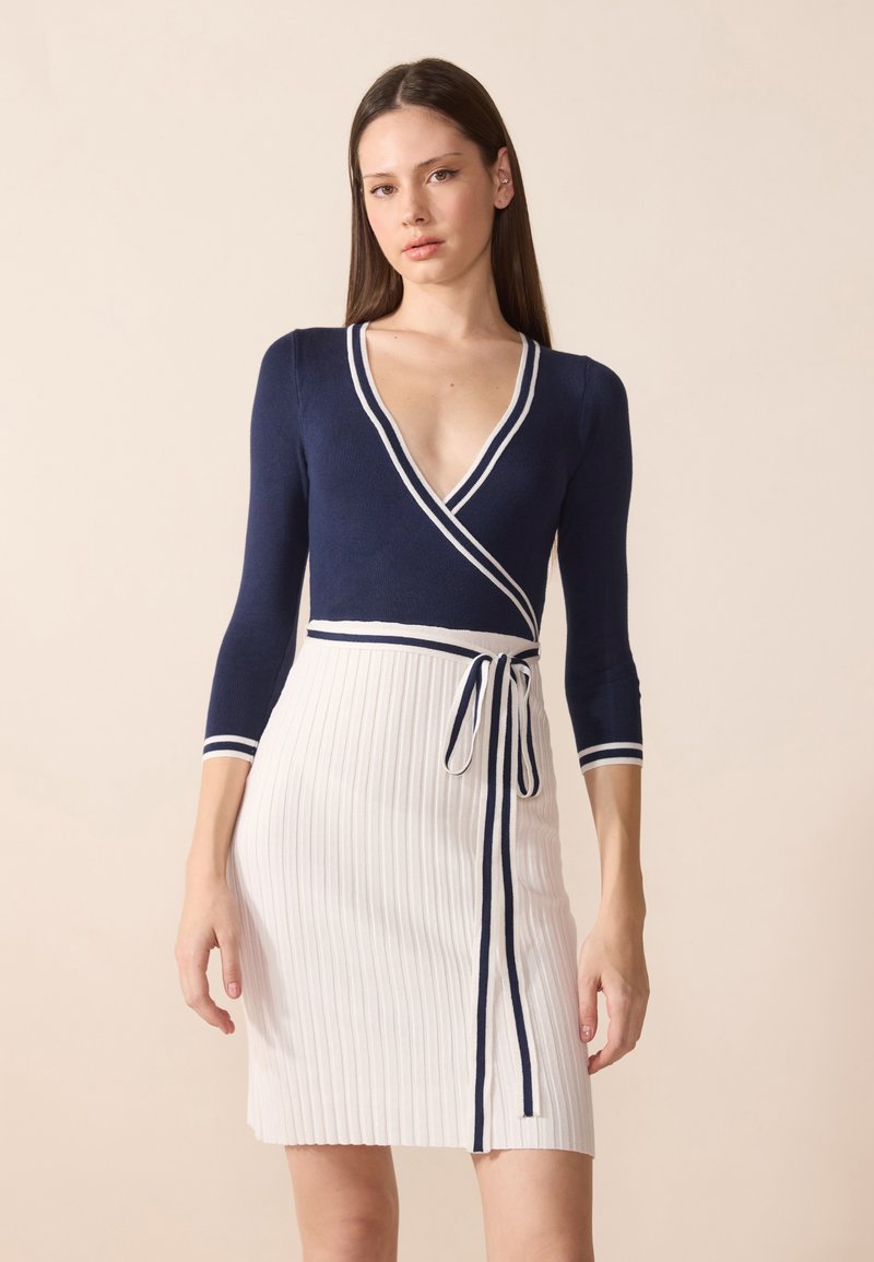 Robe portefeuille bleu marine avec des rayures blanches, décolletée en V profond, manches trois-quarts et jupe blanche côtelée avec une taille nouée.
