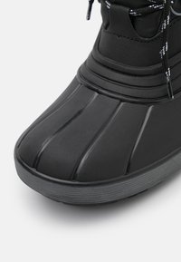 Bota impermeable negra con punta redondeada, suela de goma texturizada y un collarín acolchado en el tobillo con cordones negros y blancos.