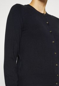 Cardigan bleu marine foncé en matériau tricoté doux. Il dispose d'un col rond, de manches longues et de cinq boutons dorés sur le devant.