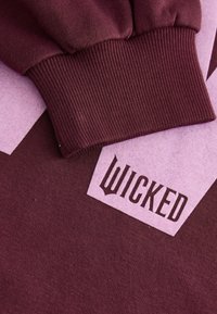Vinröd sweatshirt med ribbade muddar och en ljusrosa etikett med texten "WICKED" i mörk stil, som visar en jämn textur.