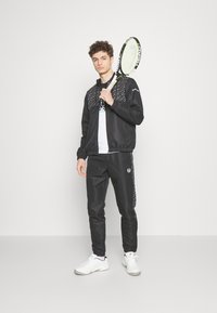 Svart idrottsjacka med mönstrade detaljer, tillsammans med svarta byxor. Modellen håller en tennisracket, klädd i en vit t-shirt och vita sneakers.