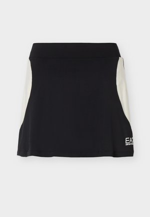 Jeune personne debout portant une chemise bleu clair à col, une jupe noire avec des panneaux latéraux clairs, des chaussettes blanches et des baskets blanches sur un fond uni.