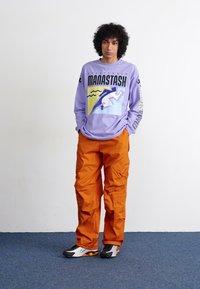 Long-sleeve purple shirt with graphic design; orange cargo pants; black and white sneakers; model standing on a blue carpet against a white wall.