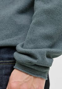 Un țesătură de pulover gri, texturată, cu un mansetă cu dungi, parțial răsucită, care dezvăluie un fundal din denim și o mână care ține țesătura.