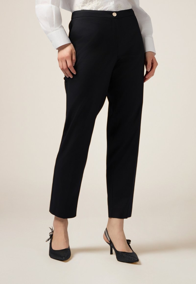 Pantaloni navy su misura con una consistenza liscia, lunghezza cropped e un piccolo dettaglio del bottone anteriore, abbinati a tacchi slingback neri.