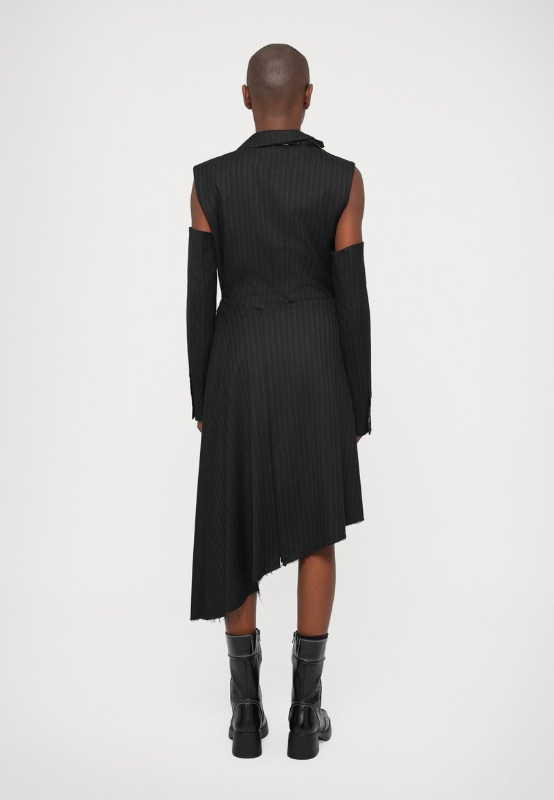 Schwarzes Nadelstreifen-Kleid mit asymmetrischem Saum, taillierten Kragen und langen Ärmeln. Der Stoff hat eine strukturierte Textur. Getragen mit schwarzen Kniestrumpf-Stiefeln.