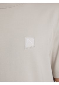 T-shirt in cotone beige con un piccolo design ricamato bianco a forma di logo triangolare nella parte sinistra del petto.