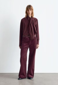 Burgundy long-sleeve shirt with neck tie paired with matching corduroy wide-leg pants; textured fabric and belt detail. Black footwear.