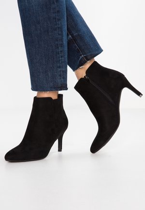 Botas de ante negras hasta el tobillo con punta afilada, tacón de 7 cm, cremallera lateral y detalles texturizados en la parte superior. Llevadas con jeans oscuros recortados.